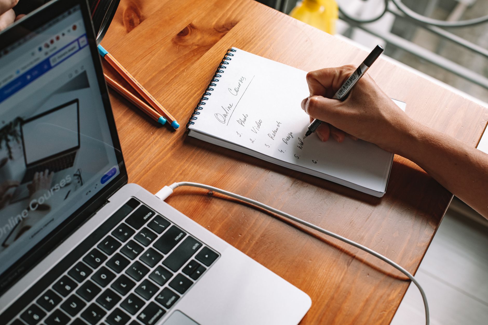 A person writes down website names on a notepad while using a laptop at a desk.